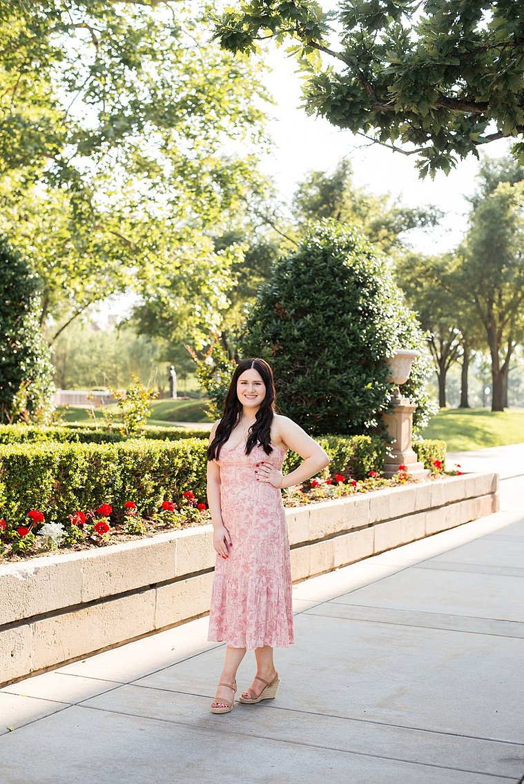 college senior girl wearing a light pink floral sundress with long dark hair stands with one hand on waist in front of a flower bed and hedges with trees around her on a sidewalk on OU campus in Norman