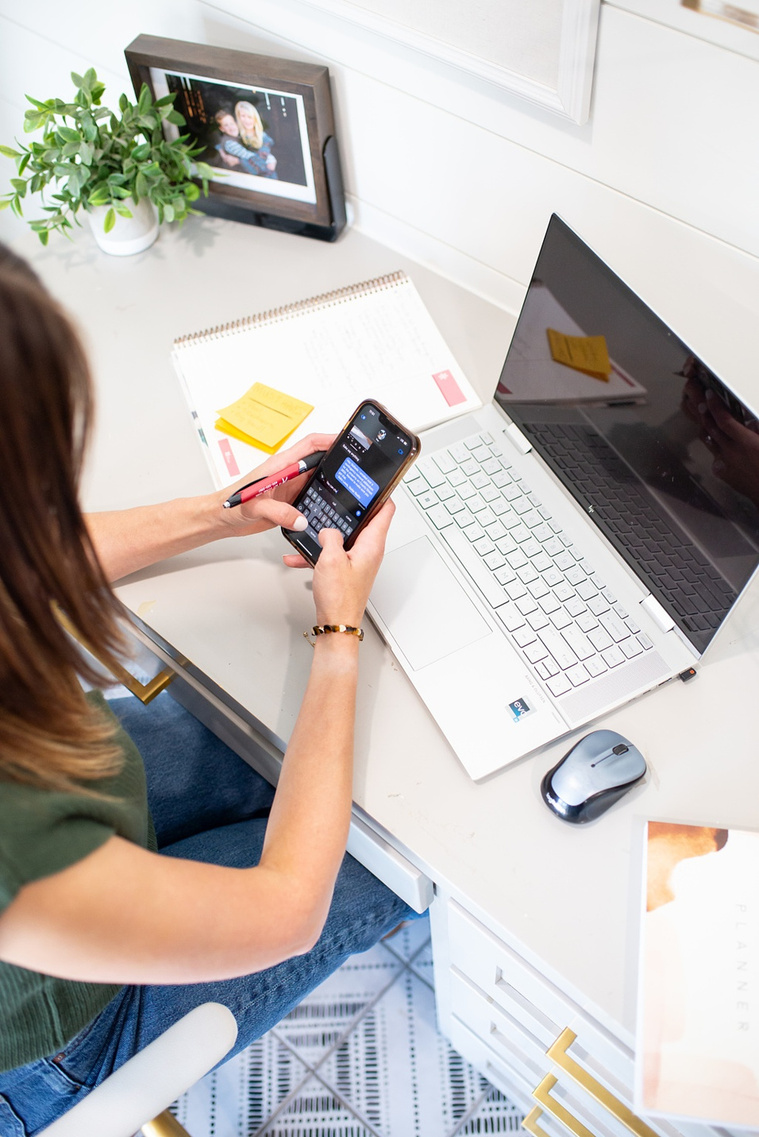 Brand photo shoot of realtor in OKC. Shot from above of her texting a client on her phone at desk with laptop, notes and family photos around her