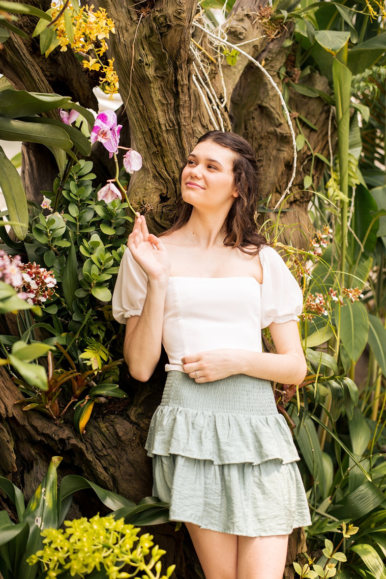 high school senior girl with long brown hair in a white blouse and green skirt holds the end of a purple flower against a tropical plant wall while she looks up dreamily inside the Crystal Bridge at Myriad Gardens in Oklahoma City 