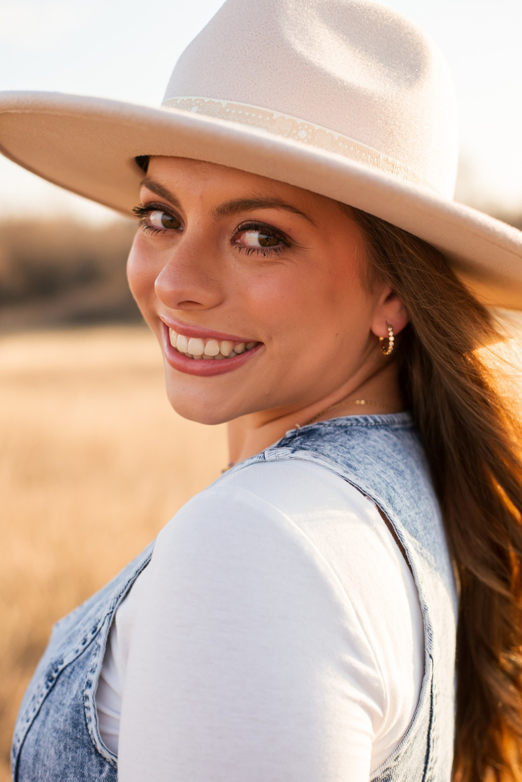 portrait of a high school senior girl with long brown hair wearing  a denim vest and western hat smiling over her shoulder as she walks through a field of golden grass in the country near OKC