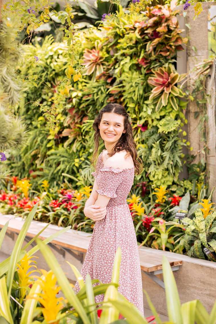 high school senior girl with long brown hair in a red and white dress stands in front of a plant wall smiling with her arms crossed and smiling over her shoulder inside the Crystal Bridge at Myriad Gardens in Oklahoma City 