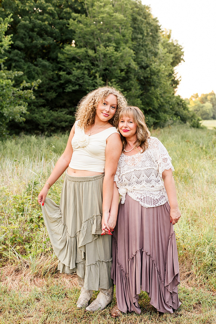 a mom and teen daughter wearing boho country outfits stand holding hands with a hand on their skirts, leaning in smiling softly in front of a grassy field and treeline in Norman, Oklahoma.