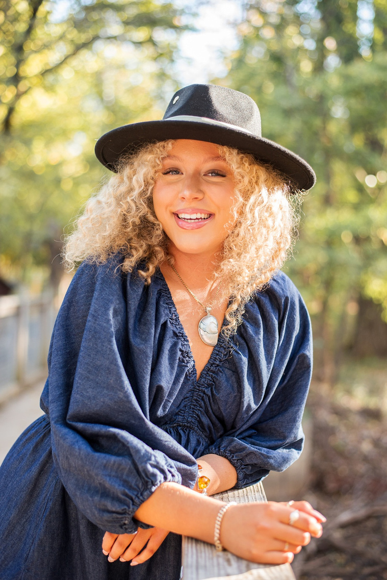 portrait of a high school senior girl with blonde curly hair and blue eyes wearing a black western hat and denim dress leaning against a bridge and laughing at Martin Nature Park in Oklahoma City