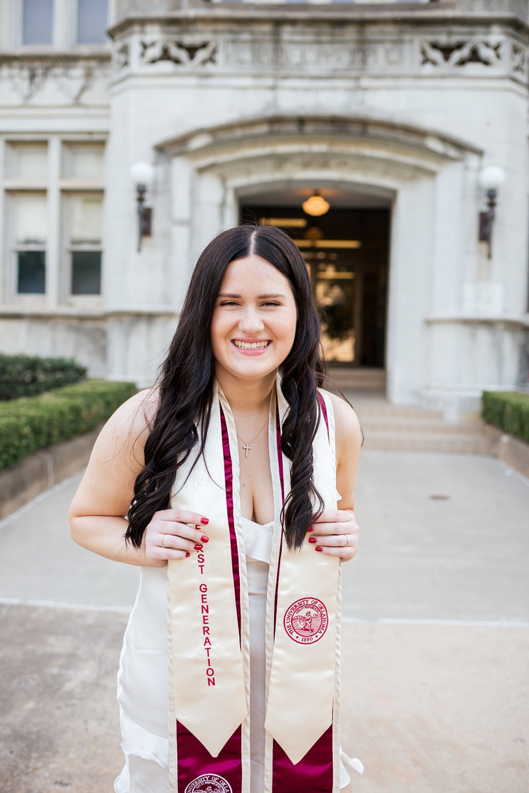 college senior girl wearing white dress and graduation stole grabs both edges of stole and is laughing while she stands on a sidewalk in the Parrington Oval on the OU campus in Norman with the Chemistry building behind her