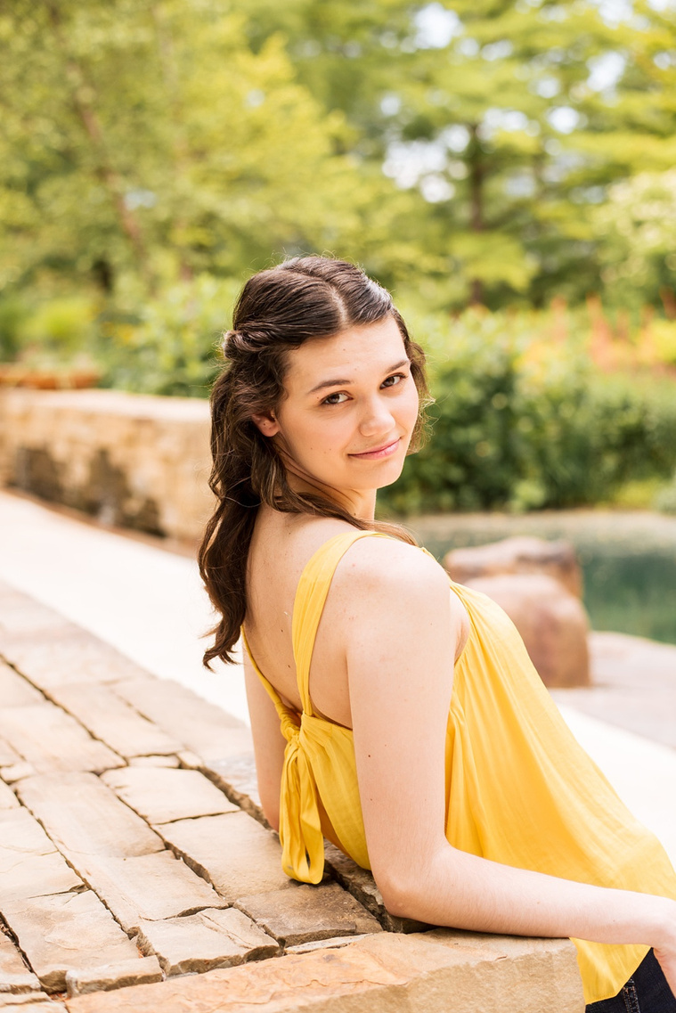 high school senior girl with long brown hair wearing a yellow top and jeans leans back on her elbows  and smiles over her shoulder at Myriad Gardens in Oklahoma City