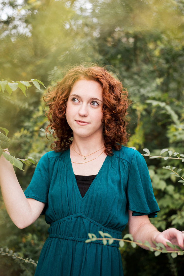 senior girl with red hair wearing teal dress stands in the midst of green foliage and looks off softly at Sutton Park in Norman, Oklahoma