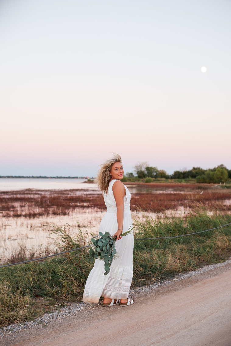 vertical portrait of a high school senior girl in a white flowy dress holding a bouquet of flowers as she walks along the road at Lake Hefner in Oklahoma City with a full  moon behind her