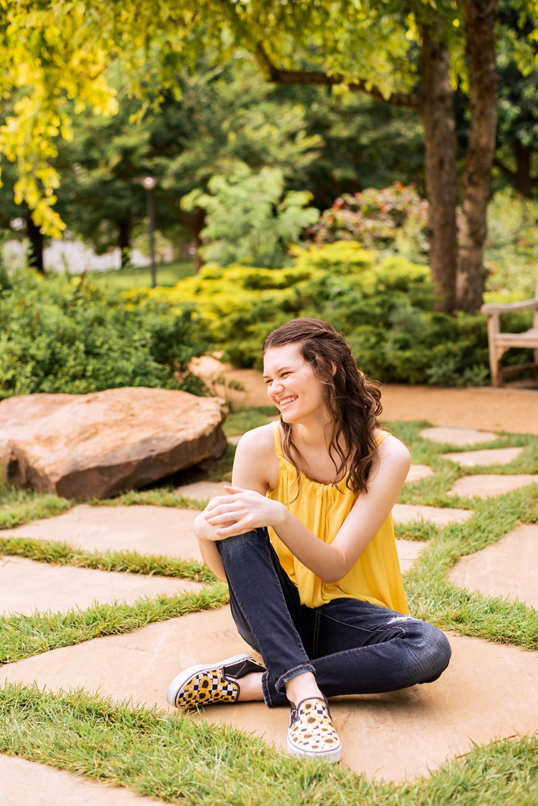 high school senior girl with long brown hair wearing a yellow top and jeans sits on a rock path with trees and plants behind her holding up on knee and laughing to the side at Myriad Gardens in Oklahoma City
