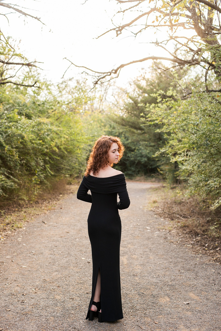 senior girl with red hair wearing an elegant long black dress poses looking over her shoulder artistically standing on a path with fall trees around her at Sutton Park in Norman, Oklahoma