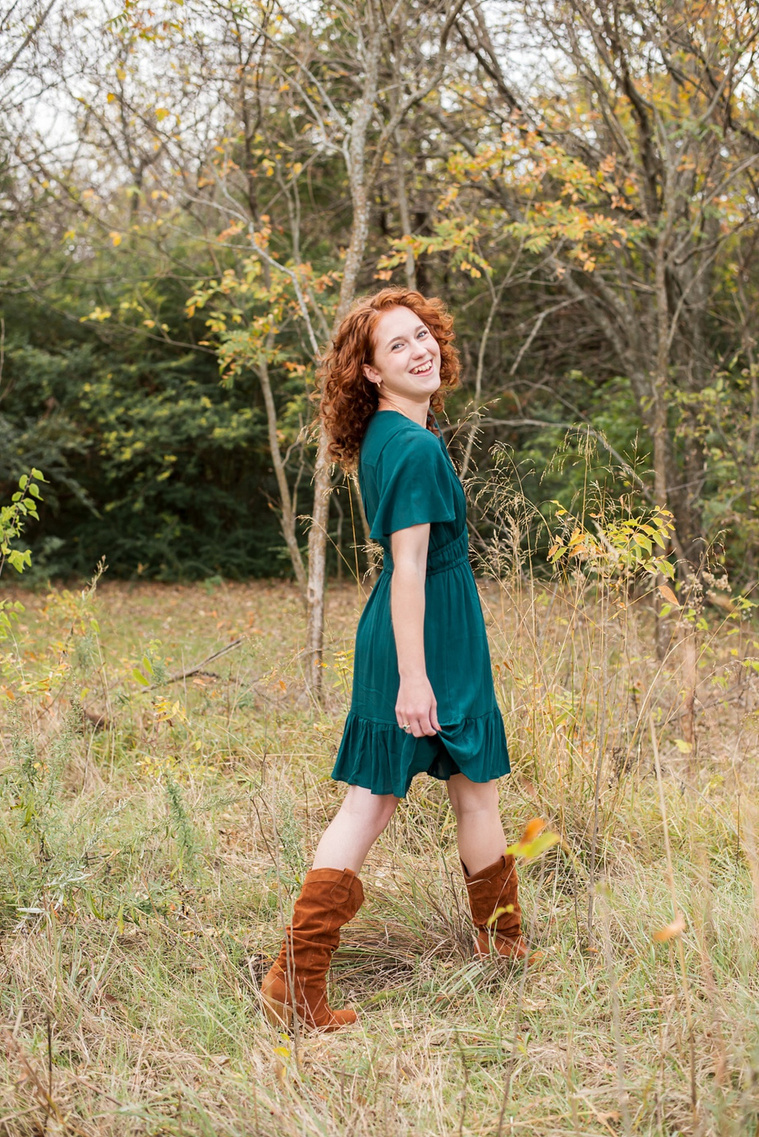 senior girl with red hair wearing teal dress steps and looks over her shoulder smiling in a field with gold grass and fall trees behind her at Sutton Park in Norman, Oklahoma