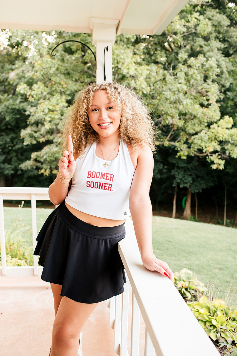 a senior girl wearing a white boomer sooner tank and black skort smiles with a number 1 finger standing on her porch in Norman, Oklahoma