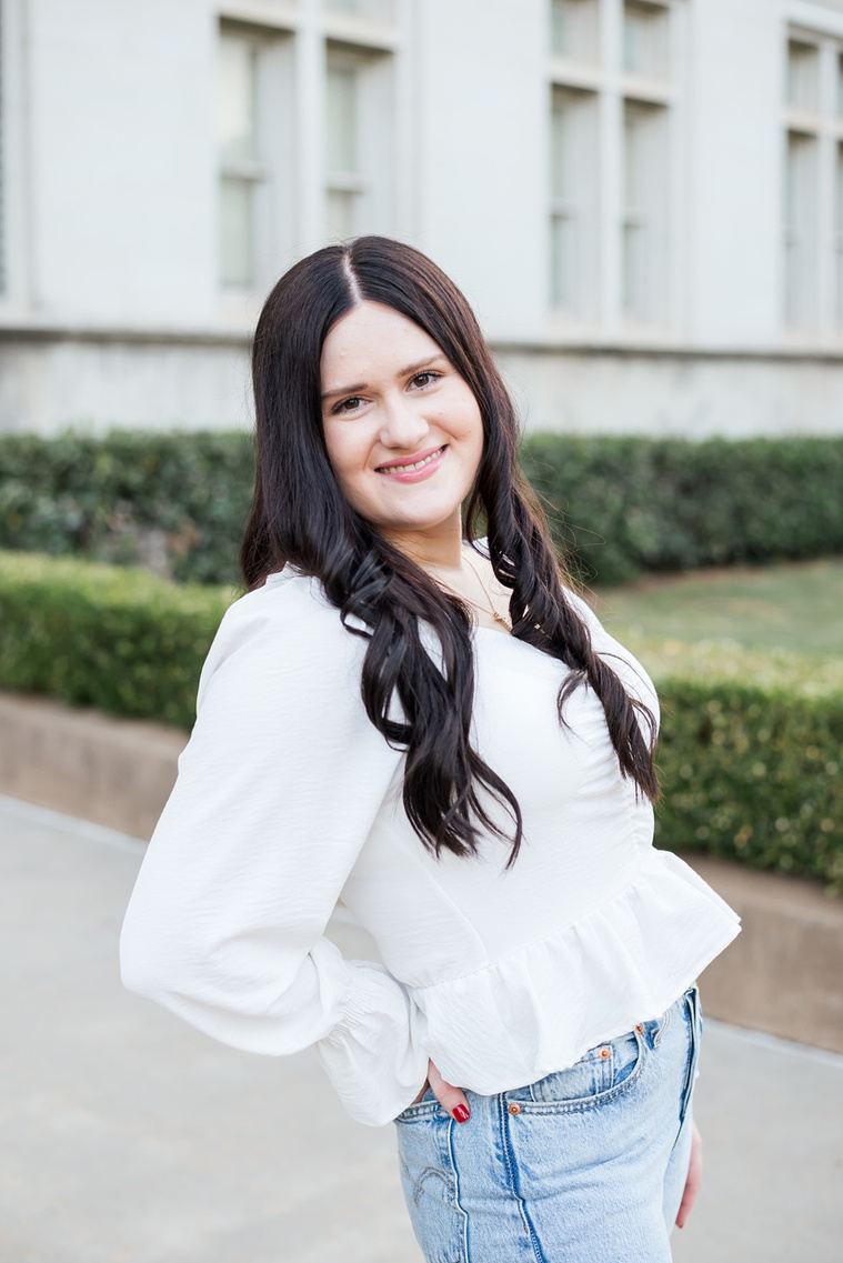portrait of a college senior girl with dark hair wearing a white blouse and jeans poses and smiles in front of the chemistry building on OU campus in Norman