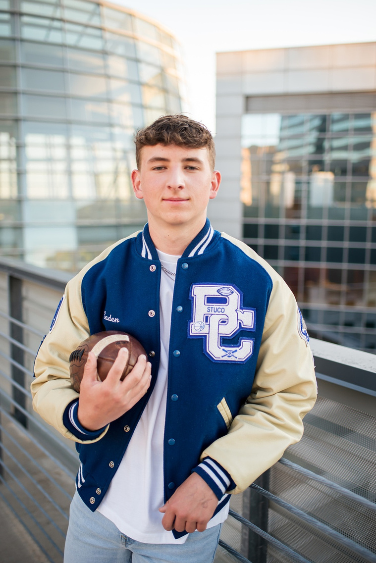 high school senior boy wearing his letterman jacket holds a football in one arm, leaning against a railing on a parking garage rooftop in downtown OKC