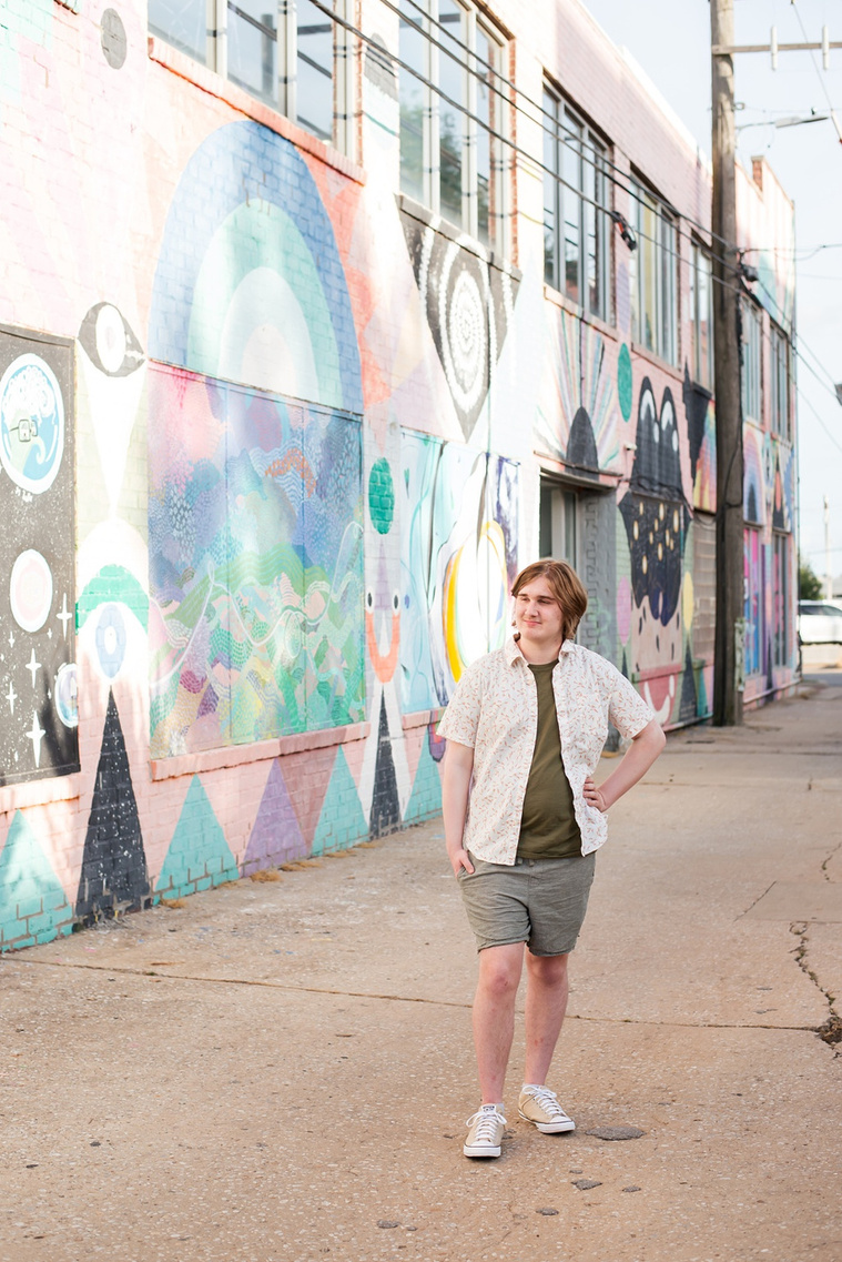 senior boy wearing a button down shirt and blue shorts stands with one hand on his hip in an alley with a colorful wall mural beside him in midtown Oklahoma City