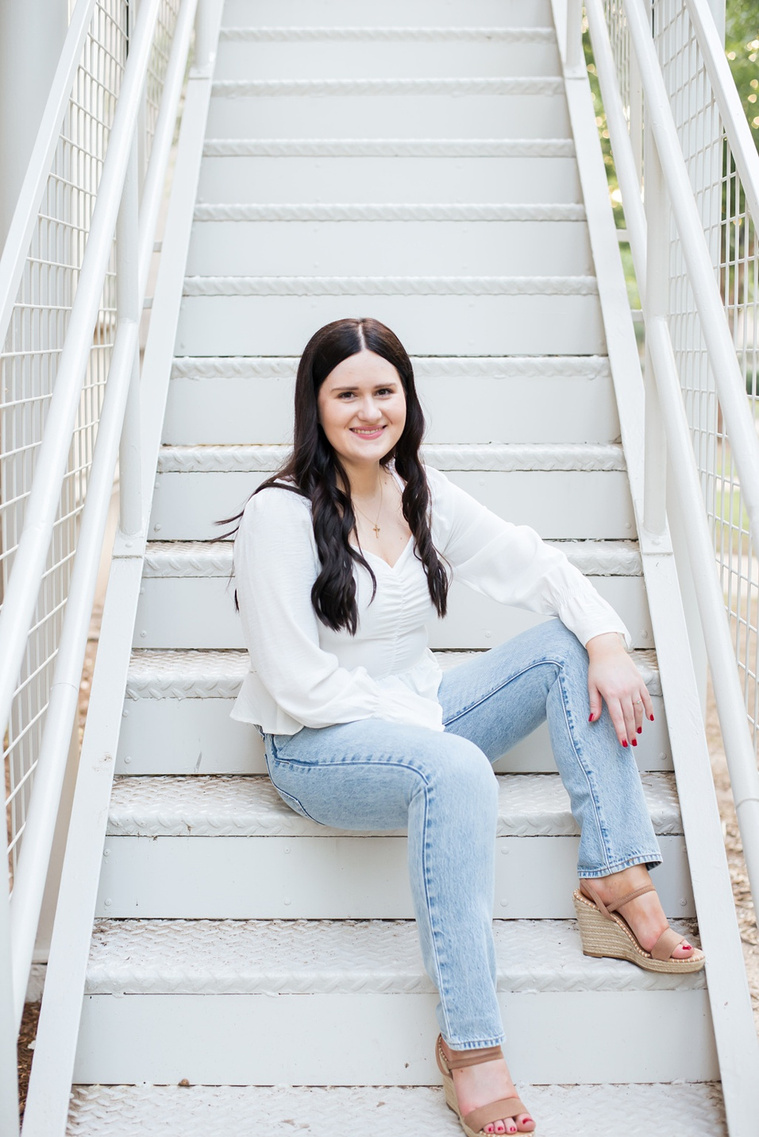 college senior girl wearing a white blouse and jeans and long dark hair sits on white steps casually and smiles by the Chemistry building on Parrington Oval on OU campus in Norman