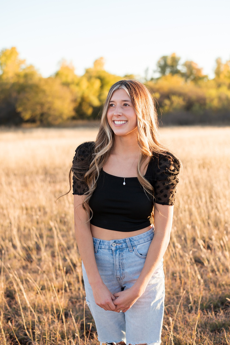 senior girl with long brown hair wearing a black top and jeans stands in tall grass with trees behind her, smiling to the side with hands in front in a country field in the fall near Oklahoma City