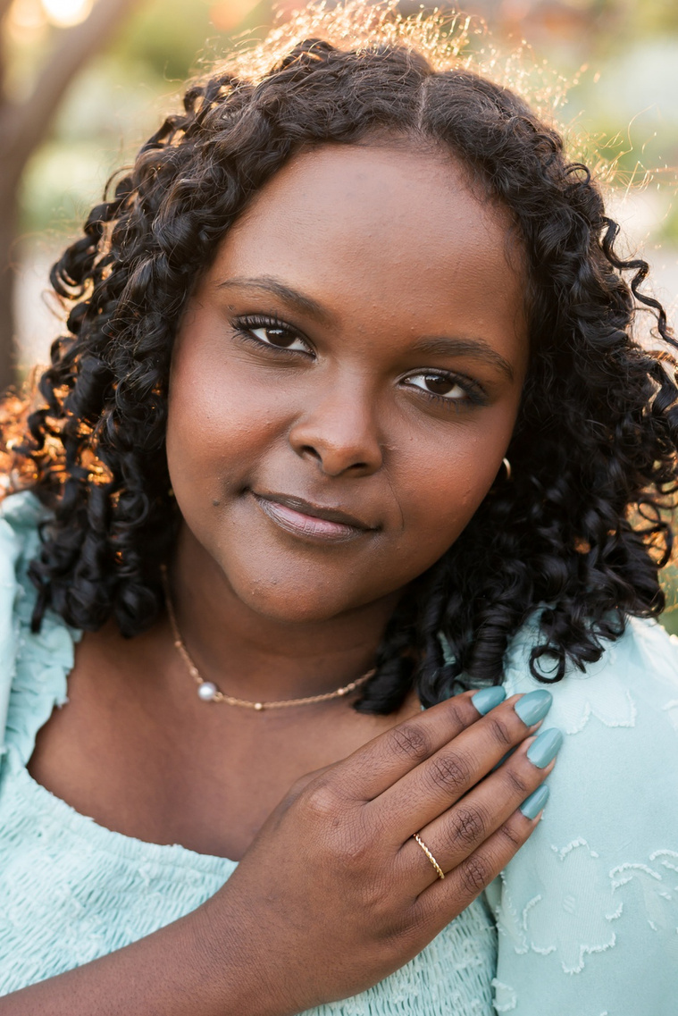 beautiful close up portrait of a black senior girl in a green dress with her hand near her face looking at camera with a soft smile and sunlight behind her