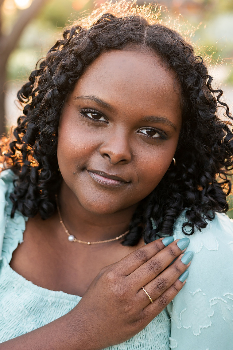beautiful close up portrait of a black senior girl in a green dress with her hand near her face looking at camera with a soft smile and sunlight behind her