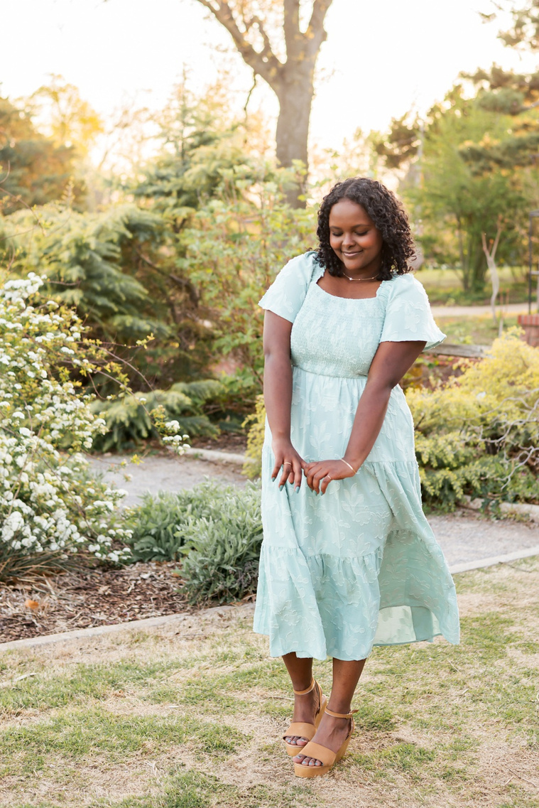 senior girl wearing a dress stands prettily and smiles shyly looking down among the flowers and trees of Will Rogers Gardens in OKC
