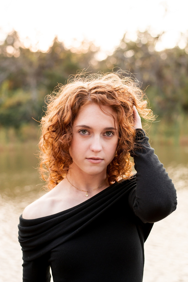 senior girl with red hair wearing an elegant long black dress stands near a pond with one hand in her hair and looking at camera with no smile at Sutton Park in Norman, Oklahoma
