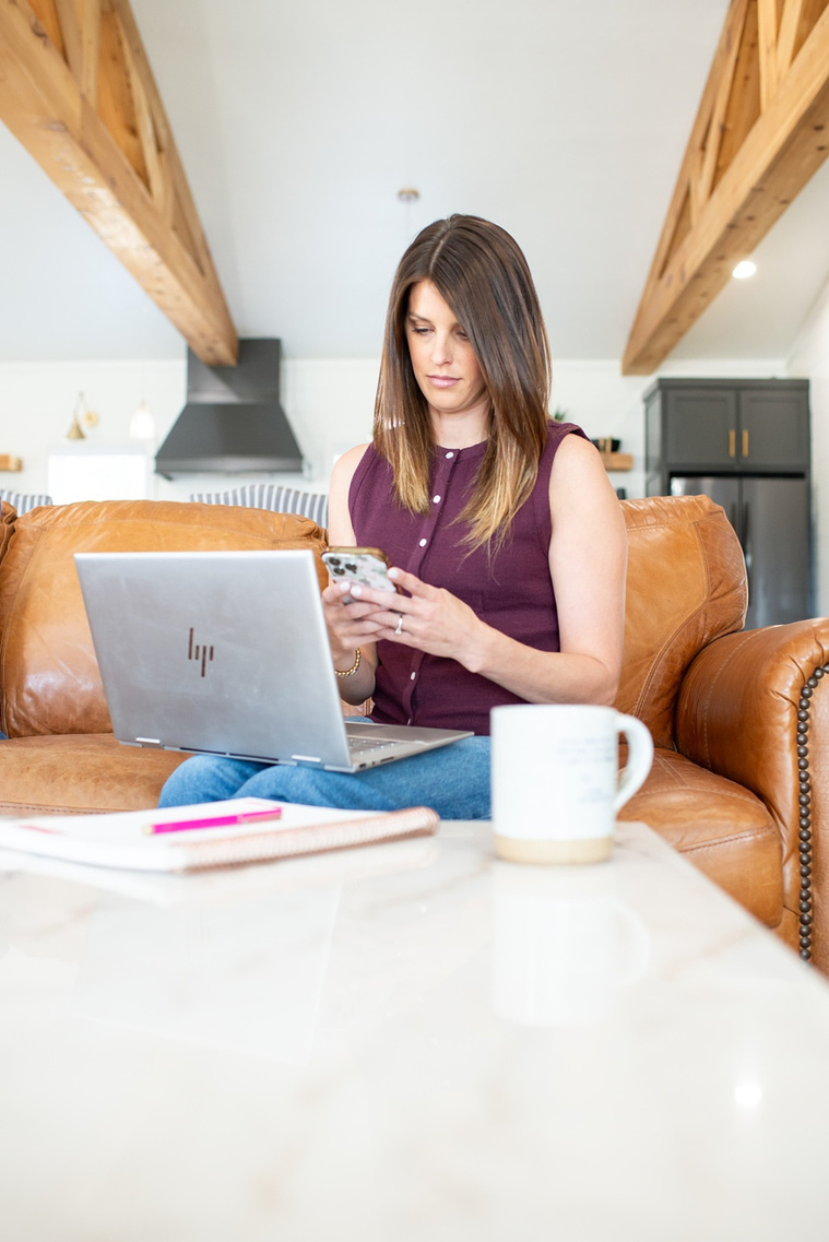 Brand photo shoot of realtor in OKC. She sits on the couch with laptop open on her lap, texting on phone with coffee mug in foreground.