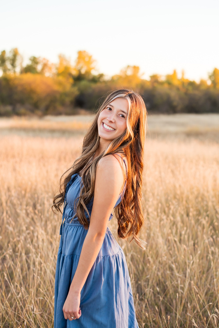 senior girl with long brown hair wearing a blue sundress smiles over her shoulder in a country field in the fall with trees behind her near Oklahoma City