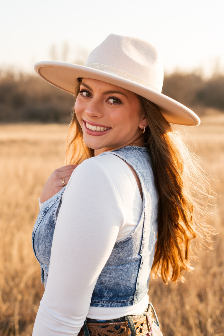 high school senior girl with long brown hair wearing Wranglers, a denim vest and western hat smiles over her shoulder beautifully as she stands in a field of golden grass in the country near OKC