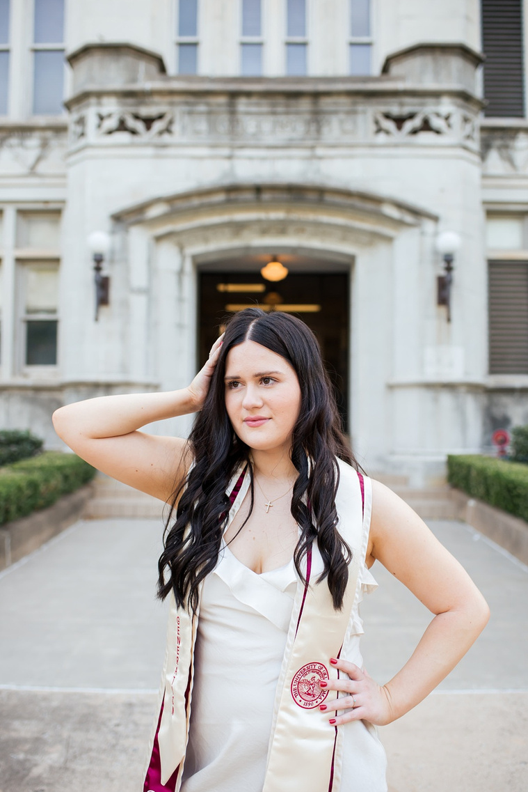 college senior girl wearing white dress and graduation stole stands on a sidewalk with one hand in hair, other on hip looking away in the Parrington Oval on the OU campus in Norman with the Chemistry building behind her