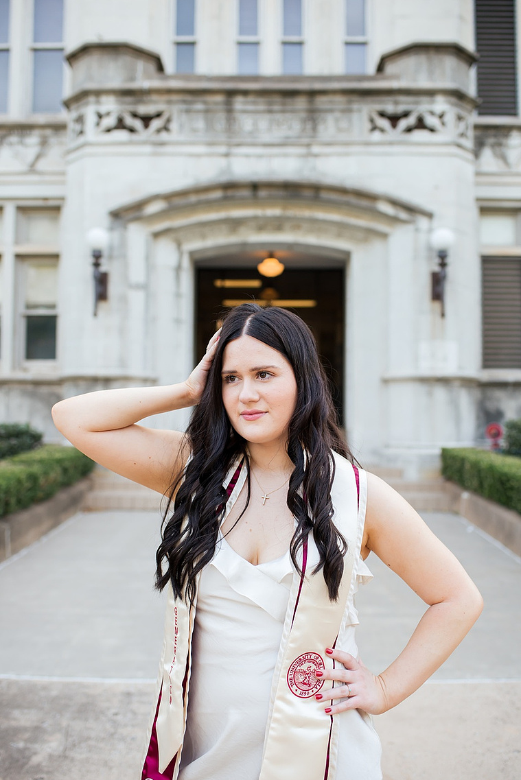 college senior girl wearing white dress and graduation stole stands on a sidewalk with one hand in hair, other on hip looking away in the Parrington Oval on the OU campus in Norman with the Chemistry building behind her
