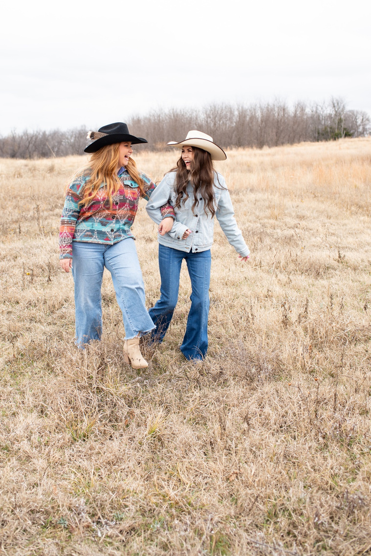 lifestyle team branding photo in OKC of two young women dressed in western clothes arm in arm walking through a field and laughing together