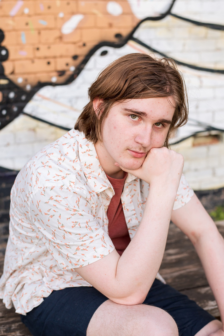 senior boy wearing a button down shirt and blue shorts sits with one hand on his chin in front of a wall mural in midtown Oklahoma City