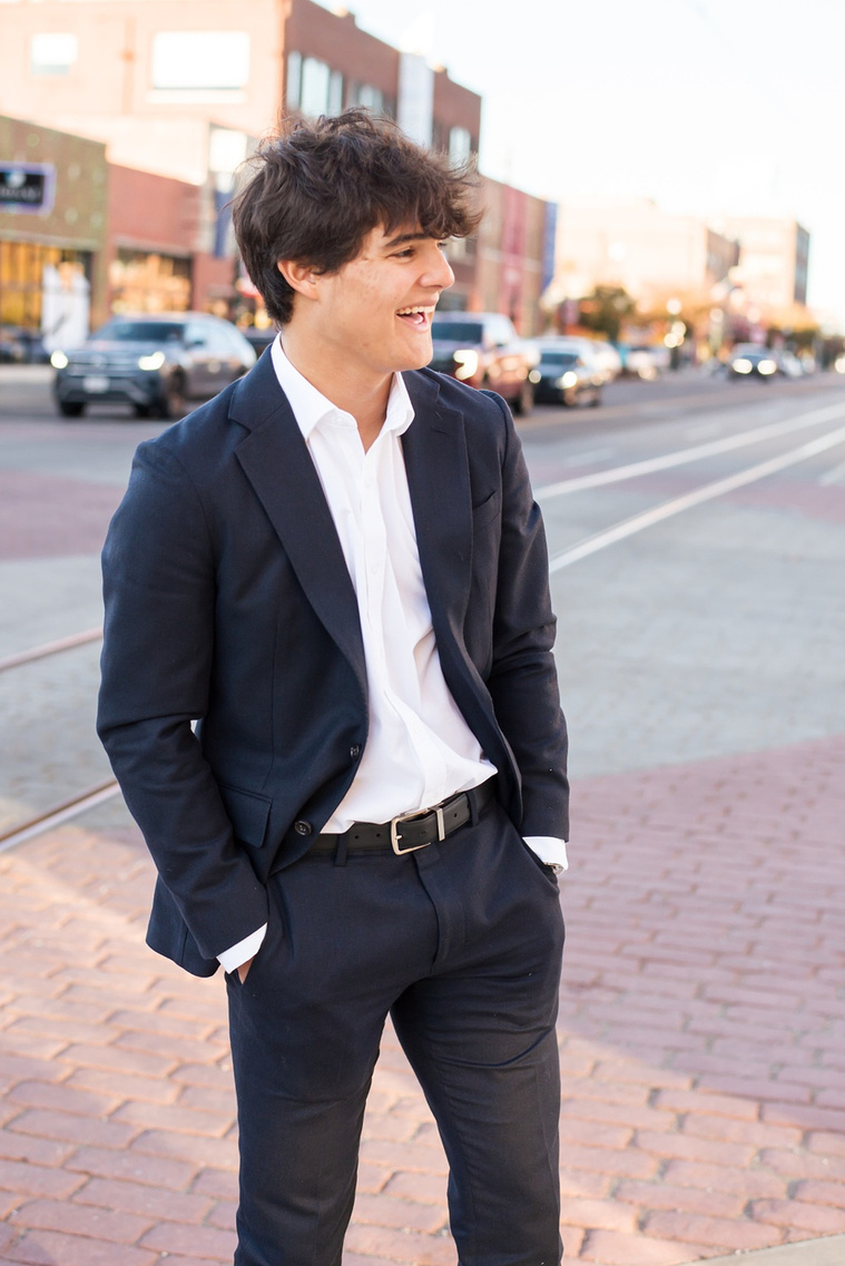a Hispanic high school senior boy wearing a dark blue suit and white shirt with his hands in his pockets looks away laughing while standing on a street corner in Automobile Alley in Oklahoma City