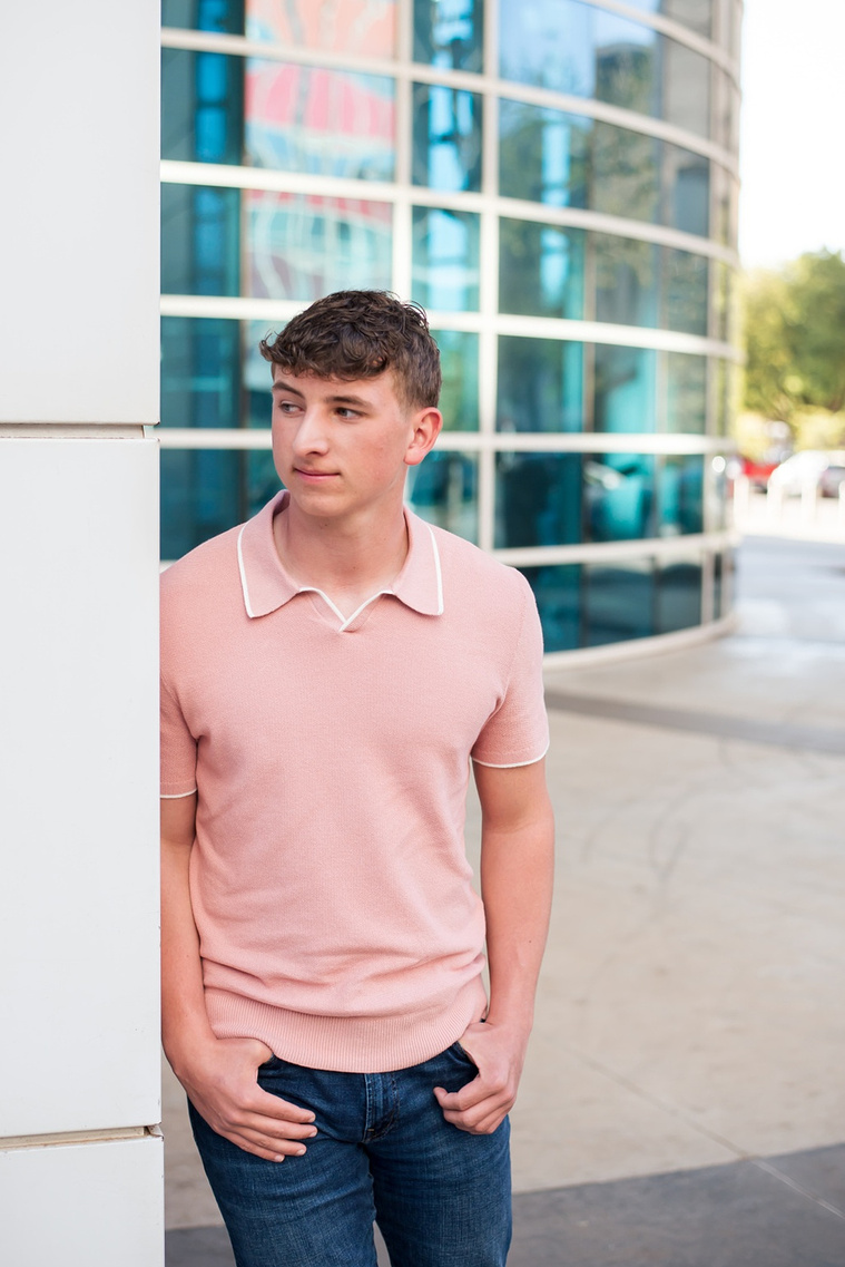 high school senior boy wearing a peach polo shirt and blue jeans stands against a white pillar looking around it in front of library in downtown OKC