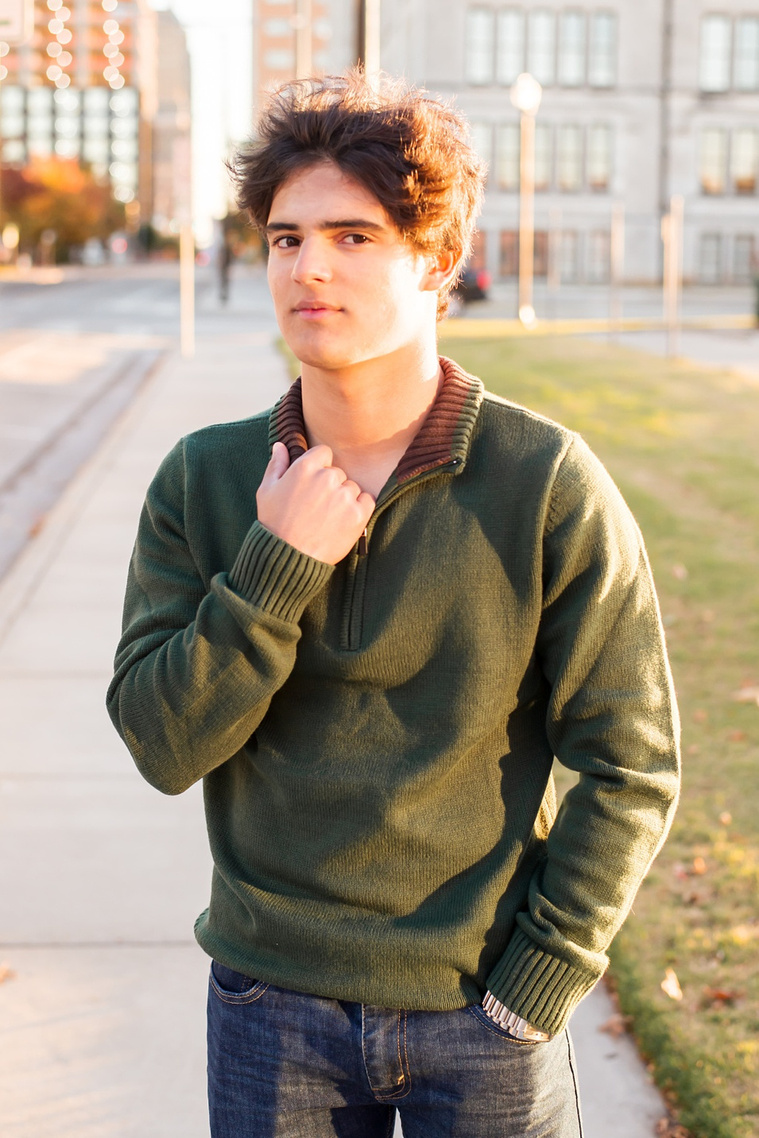 hispanic high school senior boy wearing a green sweater and jeans stands on a sidewalk in Oklahoma City with one hand in pocket and other on his collar with downtown behind him
