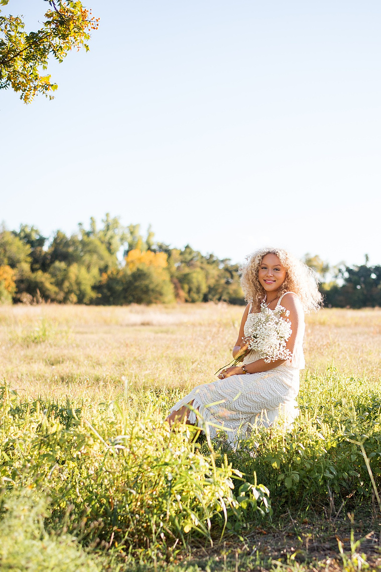 vertical portrait of a high school senior girl with blonde curly hair wearing a white fringe boho dress holding a bouquet of baby's breath sitting on a stool and smiling softly in the middle of a field at Martin Nature Park in Oklahoma City