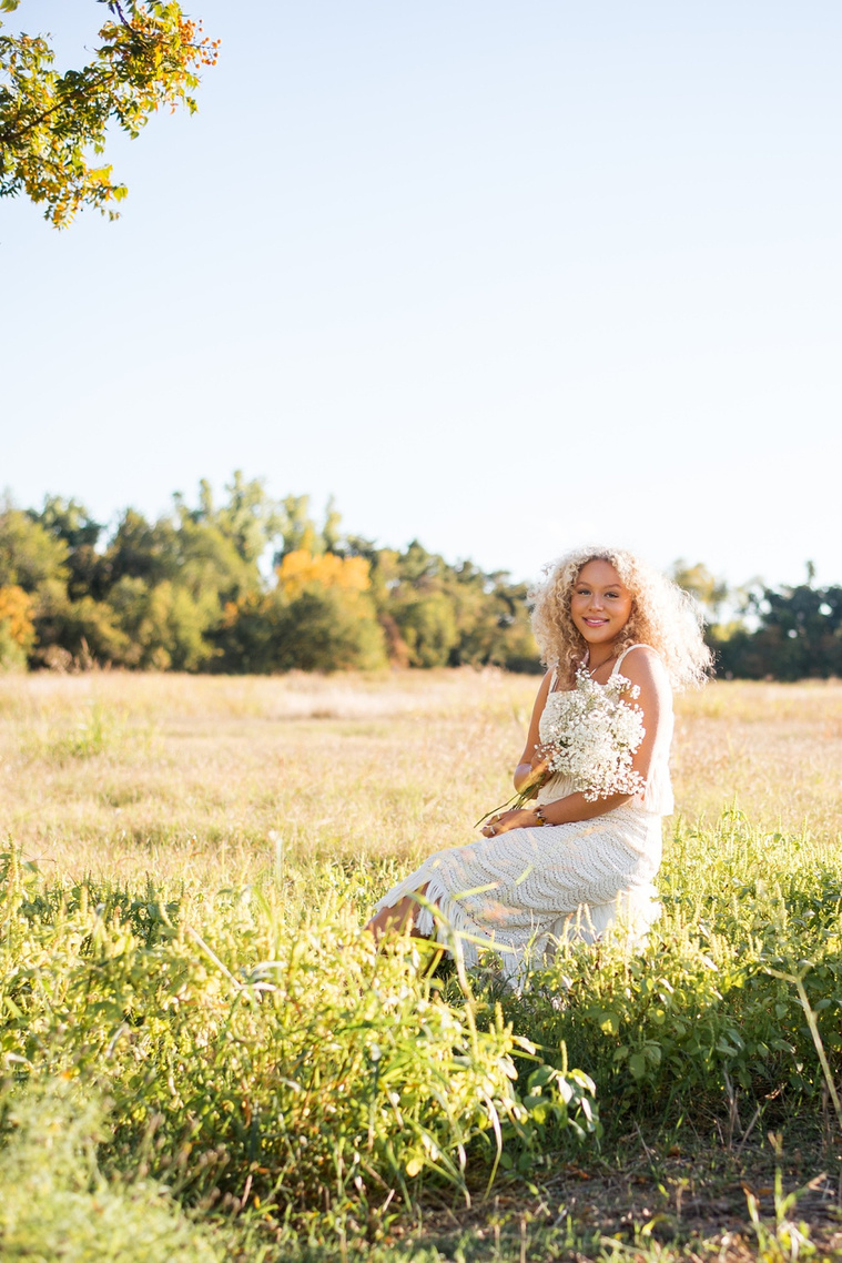 vertical portrait of a high school senior girl with blonde curly hair wearing a white fringe boho dress holding a bouquet of baby's breath sitting on a stool and smiling softly in the middle of a field at Martin Nature Park in Oklahoma City