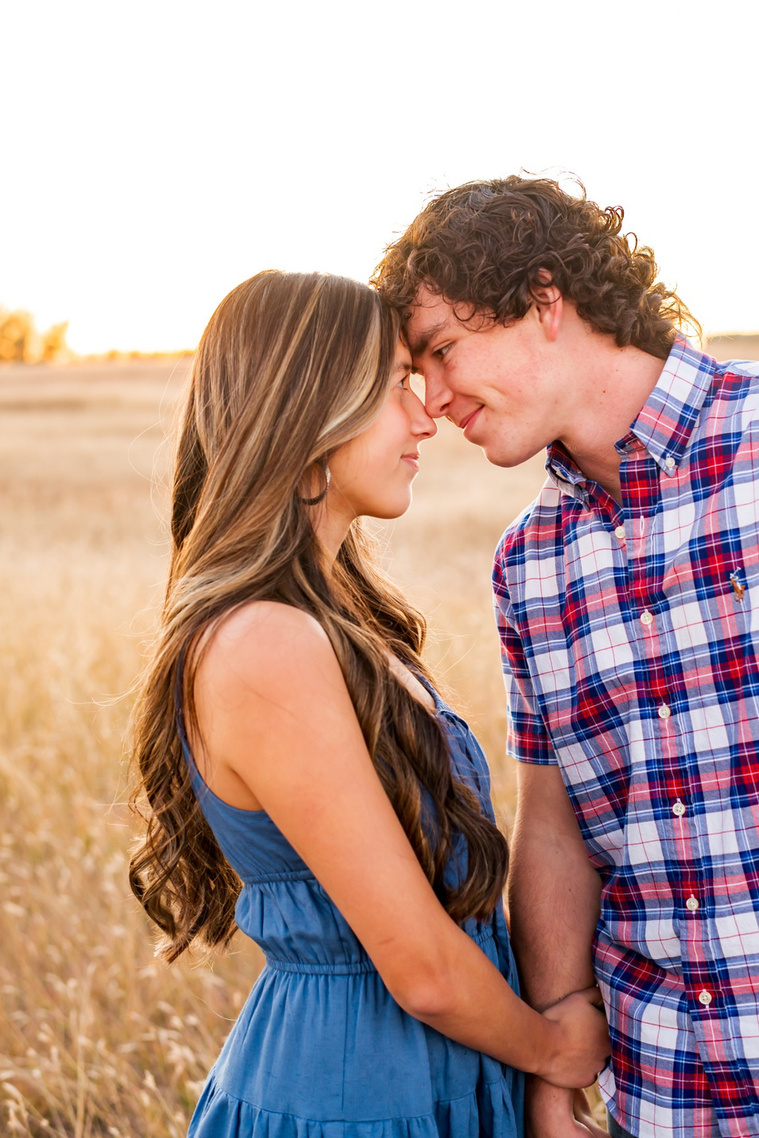 senior girl in a blue dress with her boyfriend stand together in a field in the country outside of Oklahoma City touching noses and holding hands 