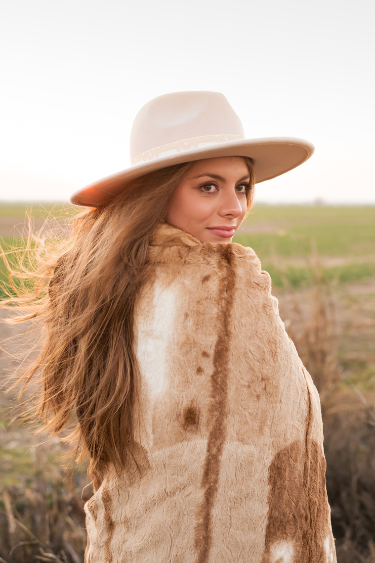 high school senior girl with long brown hair wearing a western hat wrapped in a brown blanket looks over her shoulder standing in a field near OKC with the sunset behind her