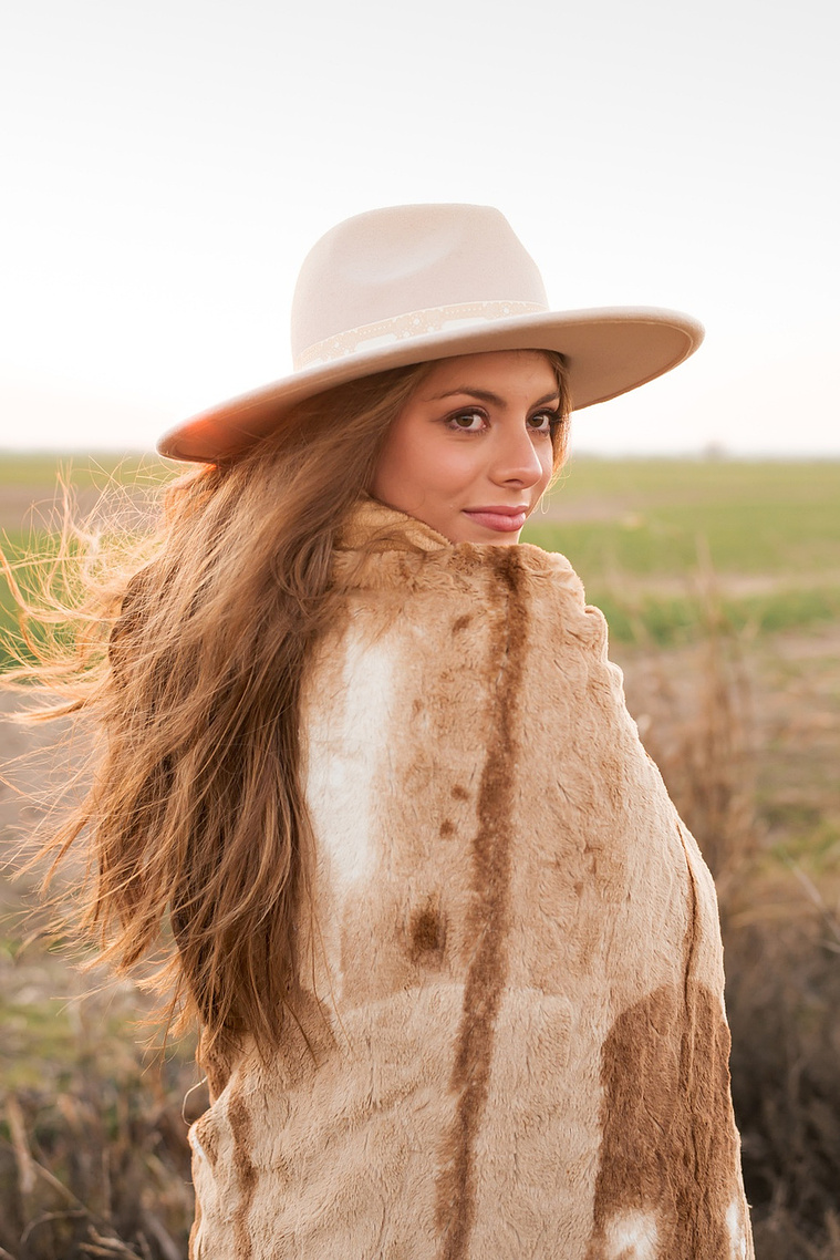high school senior girl with long brown hair wearing a western hat wrapped in a brown blanket looks over her shoulder standing in a field near OKC with the sunset behind her