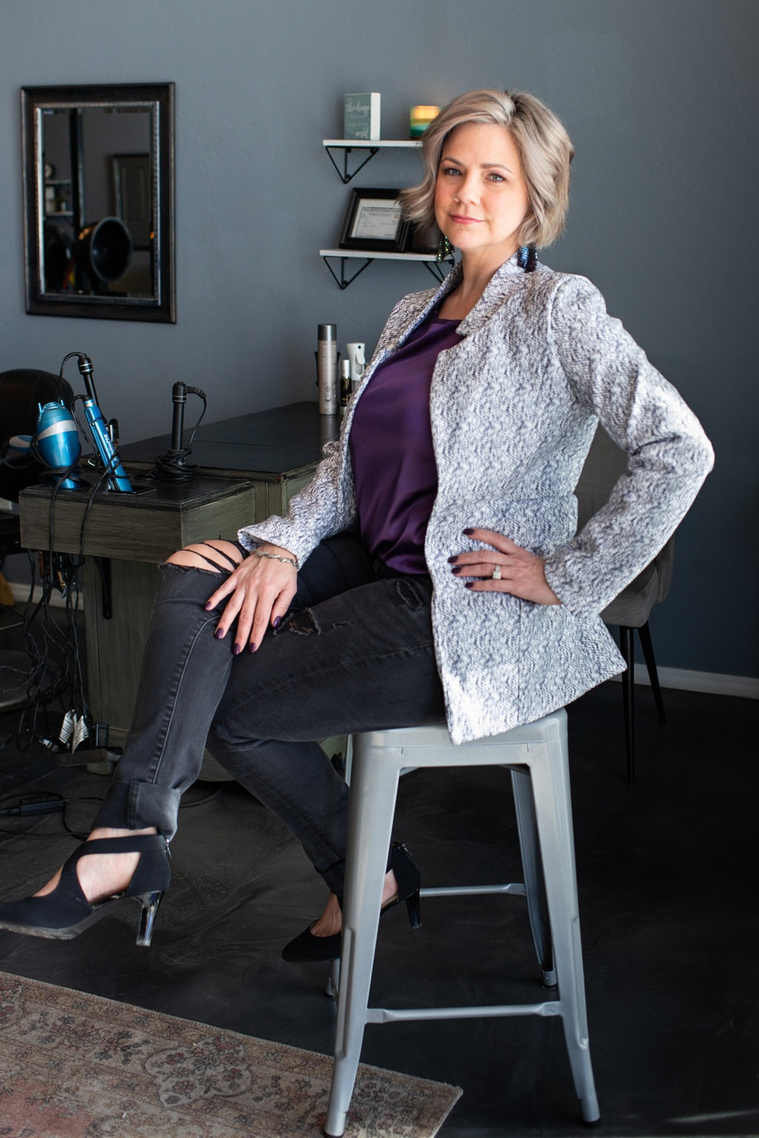Vertical portrait of a woman business owner in a gray blazer, purple top and black jeans sitting on a gray stool with her hand on hip at a branding session at Vibe Beauty Bar in Tuttle, Oklahoma