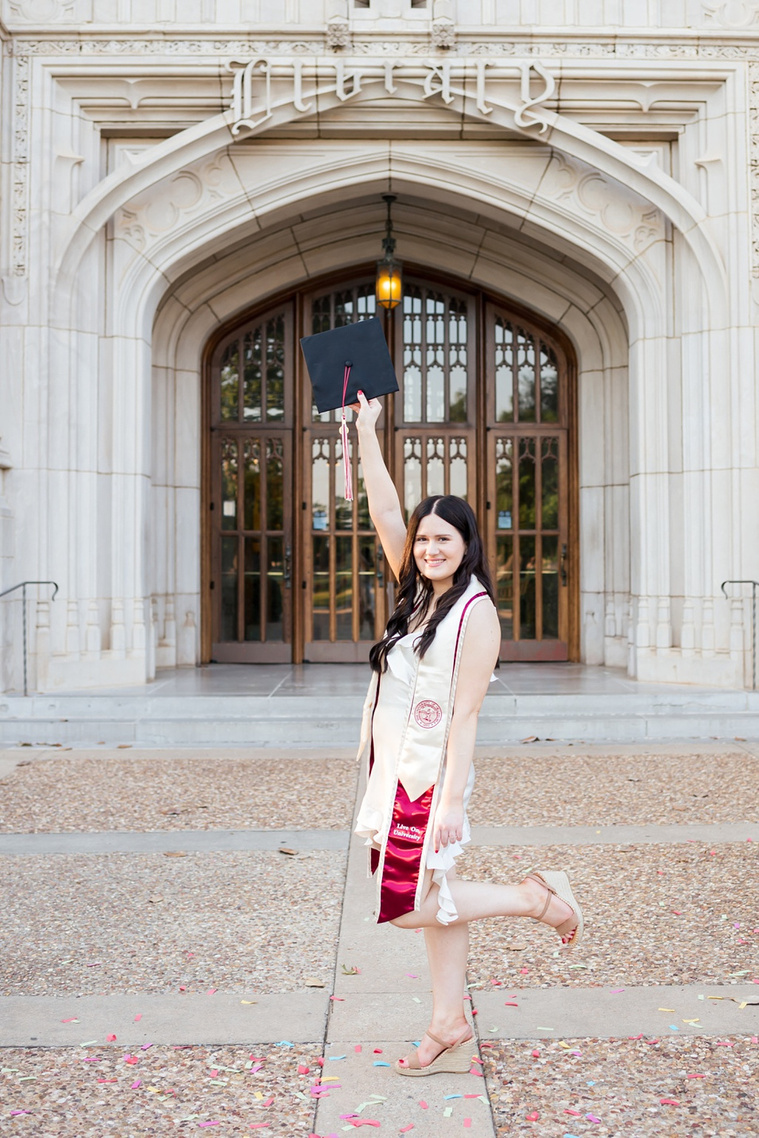 college senior girl wearing white dress and graduation stole holds up her black cap and kicks a leg out and smiles standing in front of Bizzell Library on the OU campus in Norman 