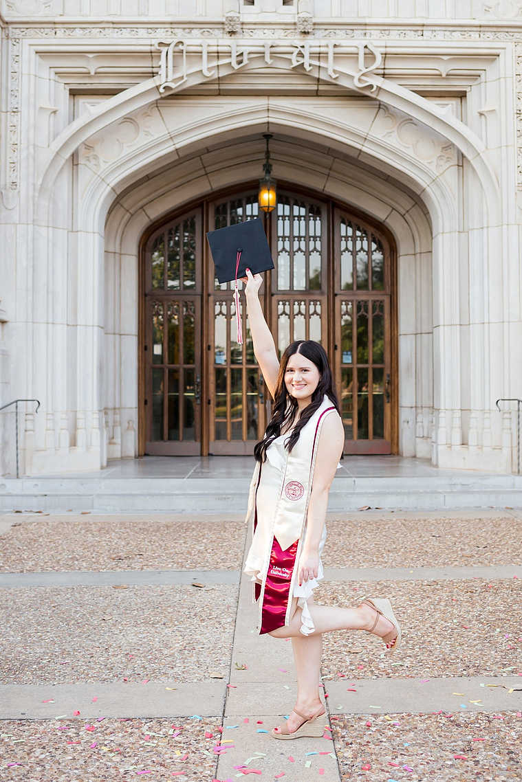 college senior girl wearing white dress and graduation stole holds up her black cap and kicks a leg out and smiles standing in front of Bizzell Library on the OU campus in Norman 