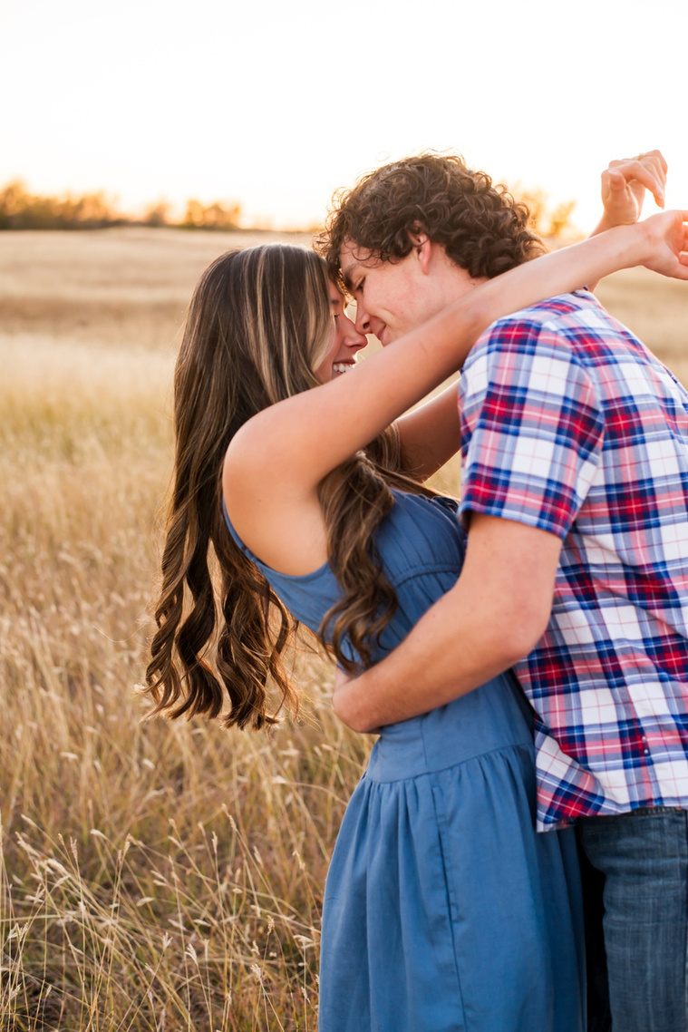 senior girl in a blue dress with her boyfriend in a plaid shirt hug each other tight and touch noses in a field in the country outside of Oklahoma City