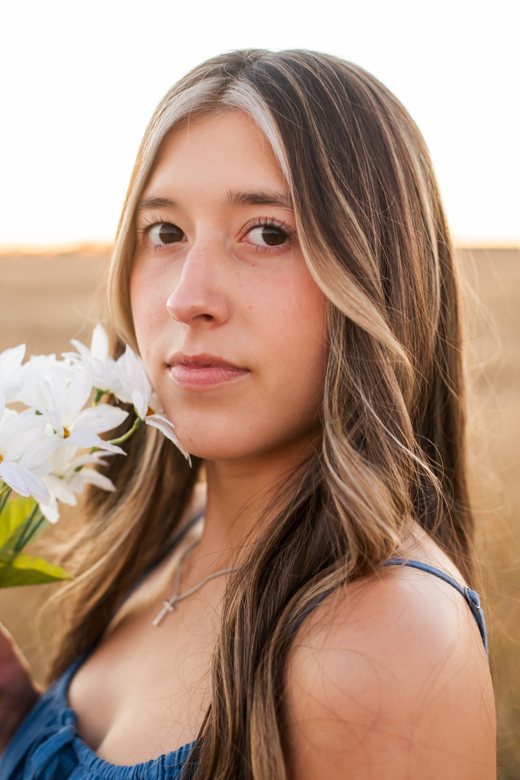 portrait of a senior girl with long brown hair wearing a blue sundress holding white daisies near her face smiling softly in a country field in the fall near Oklahoma City