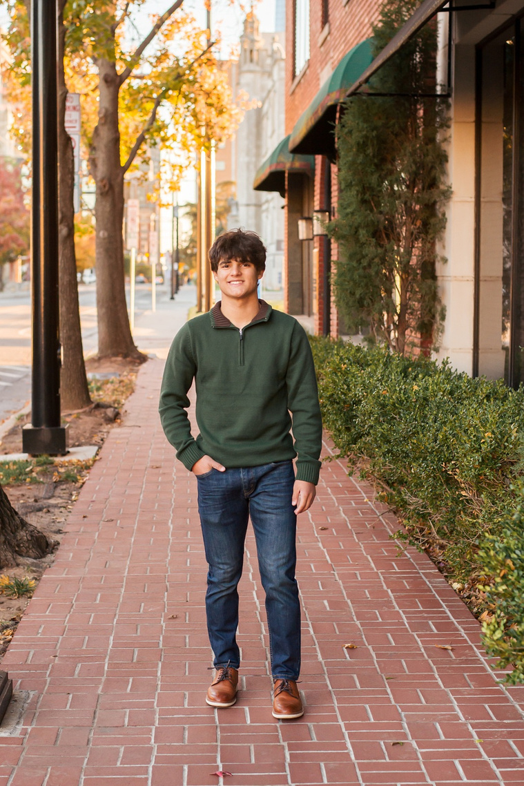 hispanic high school senior boy wearing a green sweater and jeans stands on a city sidewalk smiling with one hand in his pocket with fall trees around him and downtown Oklahoma City behind him