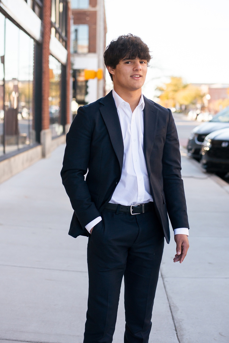 hispanic high school senior boy wearing a dark blue suit and white shirt standing on the sidewalk with a slight smile and one hand in his pocket in Automobile Alley in Oklahoma City