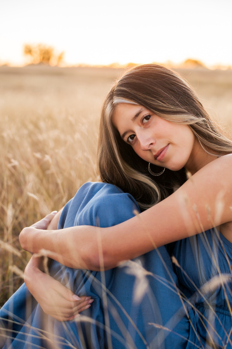 senior girl with long brown hair wearing a blue sundress sits with her knees tucked under her chin smiling softly in a country field in the fall near Oklahoma City
