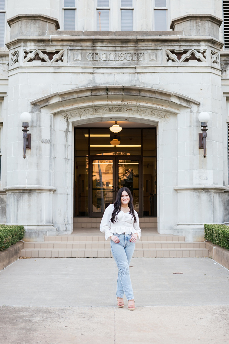 portrait of a college senior girl wearing a white blouse and jeans with long dark hair crosses her ankles with thumbs in pockets and stands in front of Chemistry building on OU campus in Norman