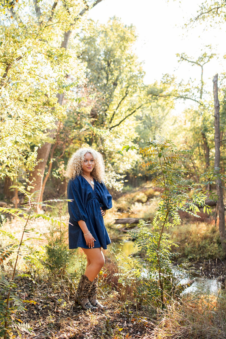 vertical portrait of a high school senior girl with blond curly hair wearing a blue dress and cowboy boots looking to the side in front of a creek and trees at Martin Nature Park in Oklahoma City