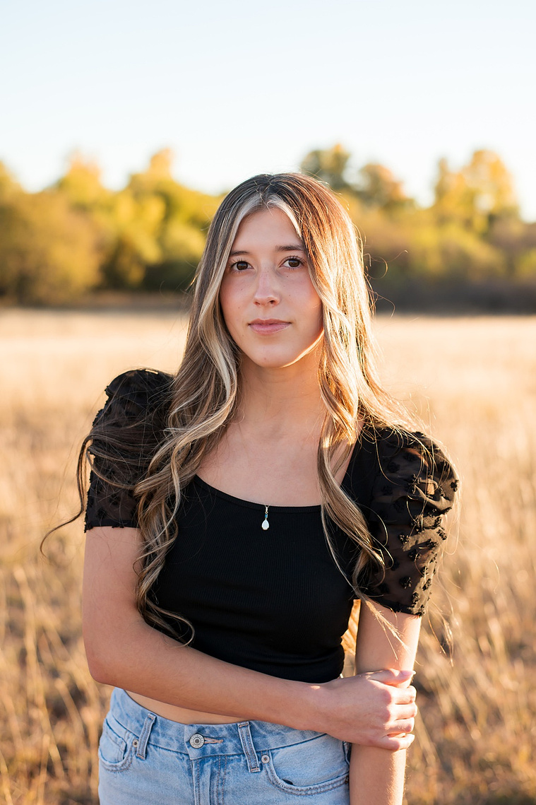 senior girl with long brown hair wearing a black top and jeans stands in tall grass with trees behind her, smiling softly in a country field in the fall near Oklahoma City